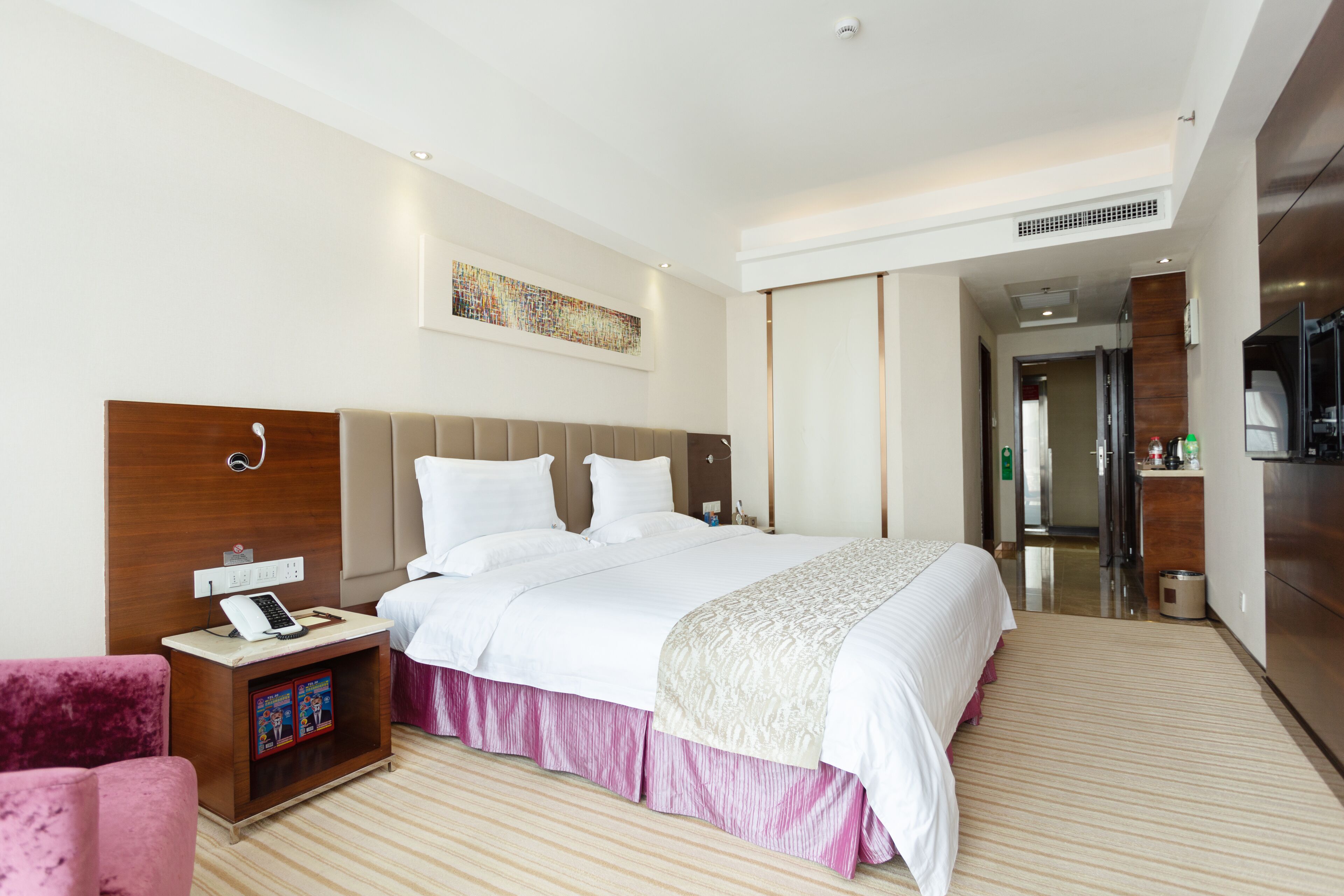Habitación de hotel con una cama grande, una mesita de noche con teléfono, una silla rosa y un televisor.