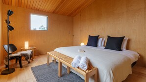 A bedroom with a wooden ceiling, a large bed with white bedding, two pillows, a bench with towels, a floor lamp, and a small table with a candle.