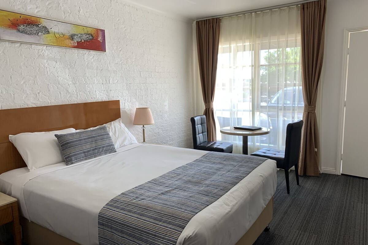 A hotel room with a bed, a chair, a small table, and a window with curtains.