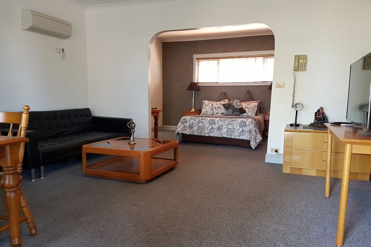 A living room with a black sofa, wooden coffee table, and a bed with patterned bedding.