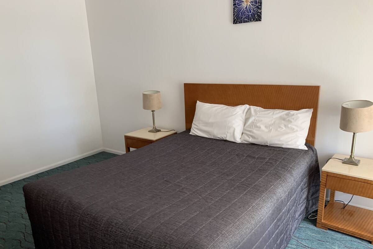 A neatly made bed with a dark grey quilt, two bedside tables with lamps, and a plain white wall with a single piece of wall art.