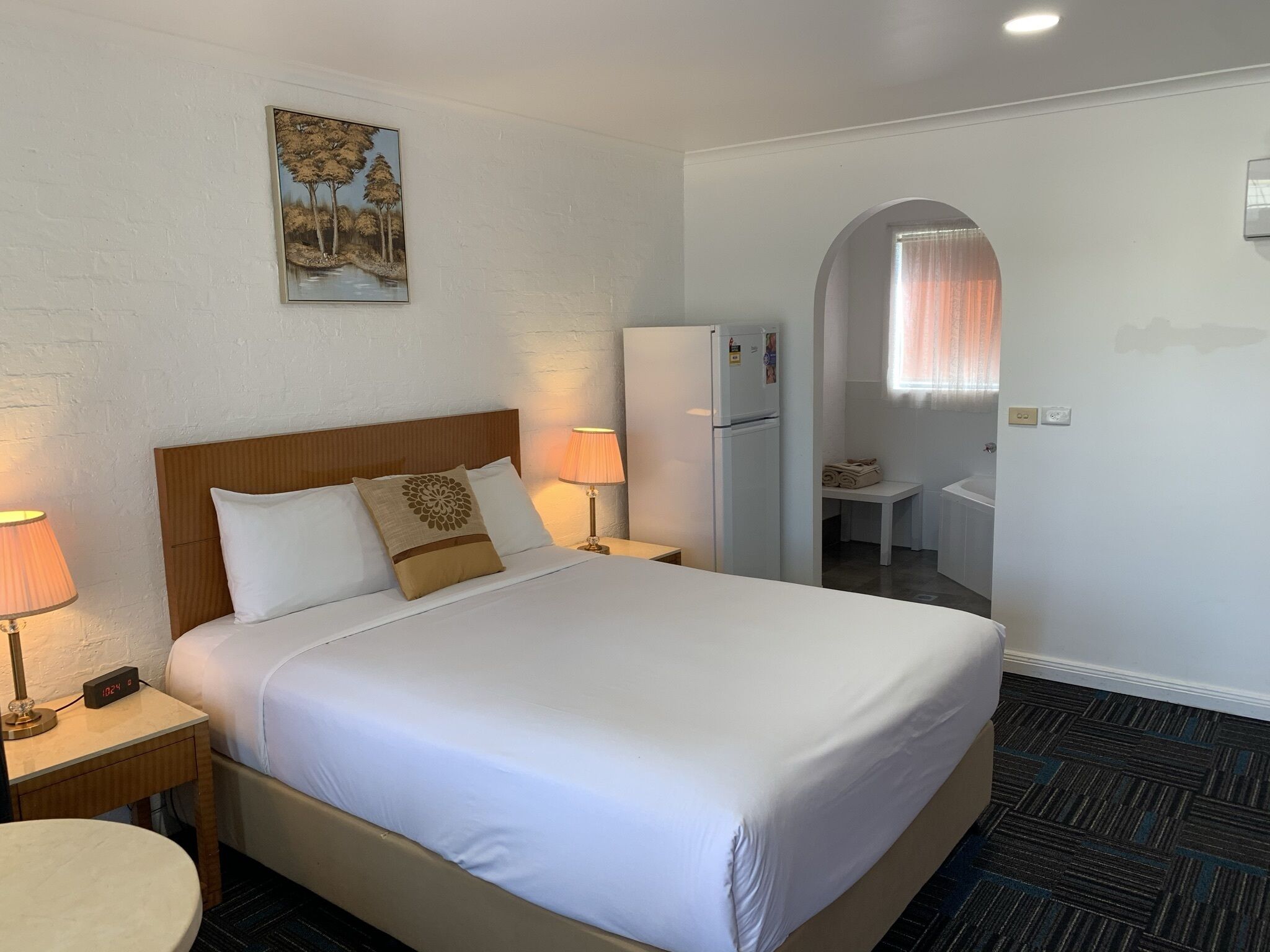 Una camera d'albergo con un letto, abat-jour, un frigorifero, un tavolino e un quadro appeso al muro.
