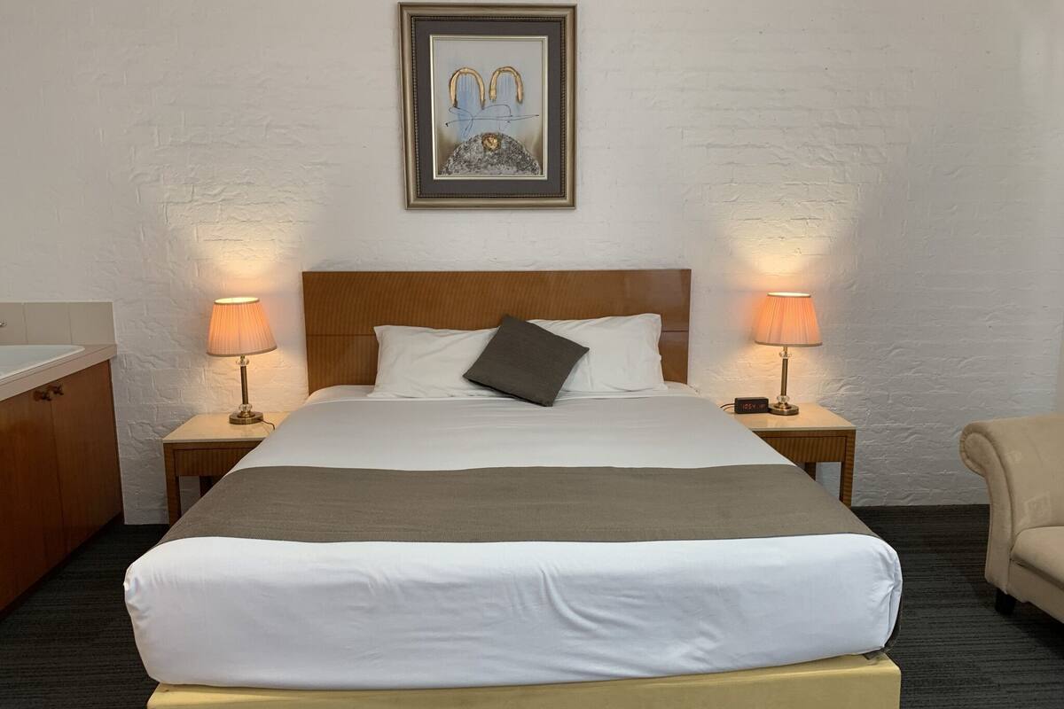 A hotel room with a large bed, two bedside lamps, a wooden headboard, a framed artwork above the bed, and a small table with a clock.