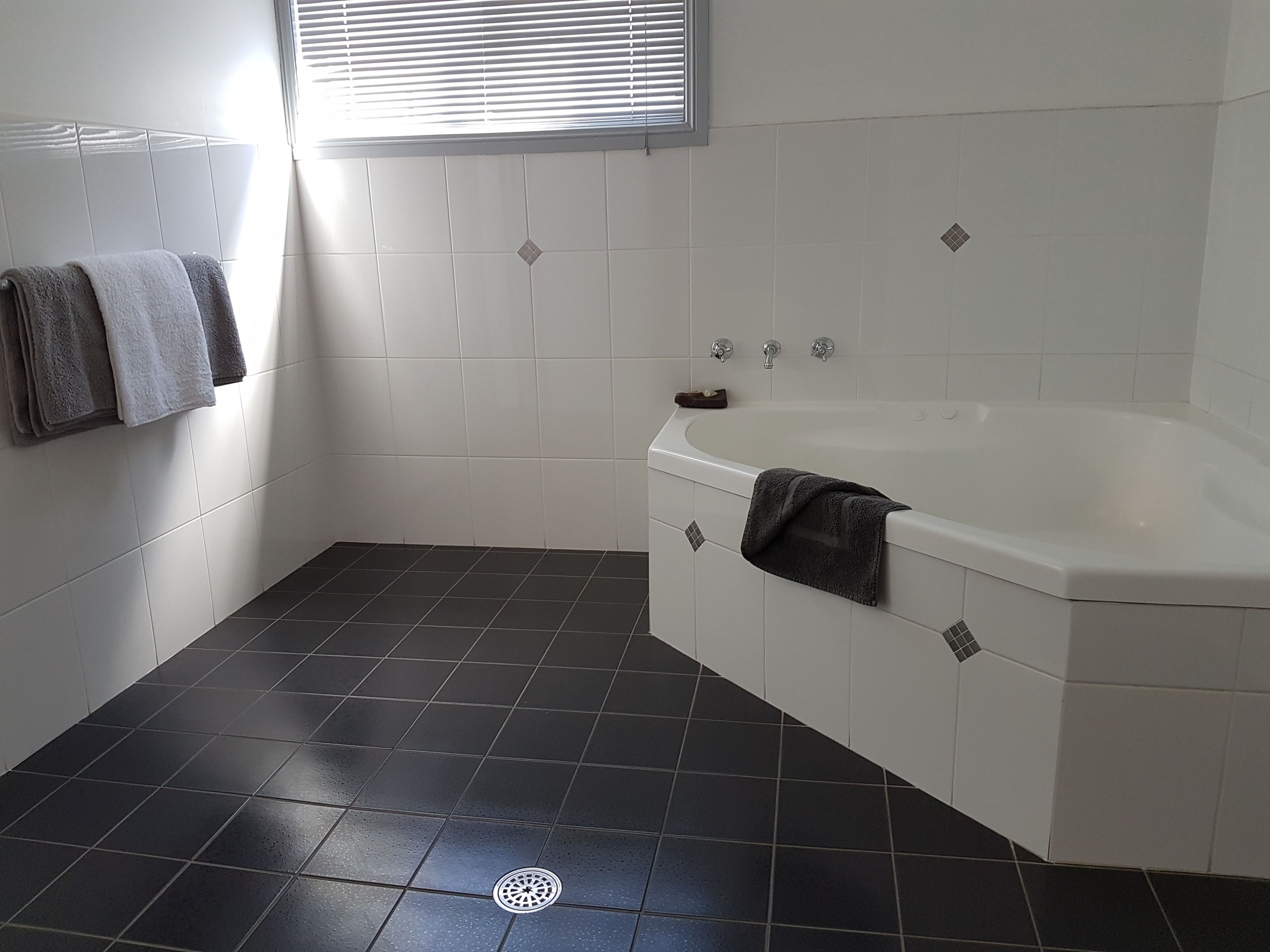 Une salle de bain avec une baignoire, des carreaux de sol foncés, des murs blancs et une fenêtre avec des stores.