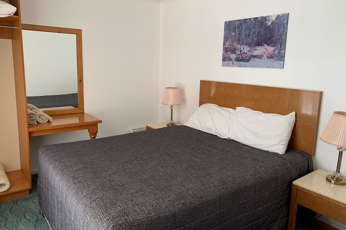 A neatly arranged hotel room with a bed, a bedside table with a lamp, a mirror, and a painting on the wall.
