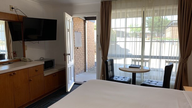 A hotel room with a bed, a TV mounted on the wall, a desk with a telephone and a laptop, a small table with a chair, and a large window with curtains. - Swaggers Motor Inn (Yass)