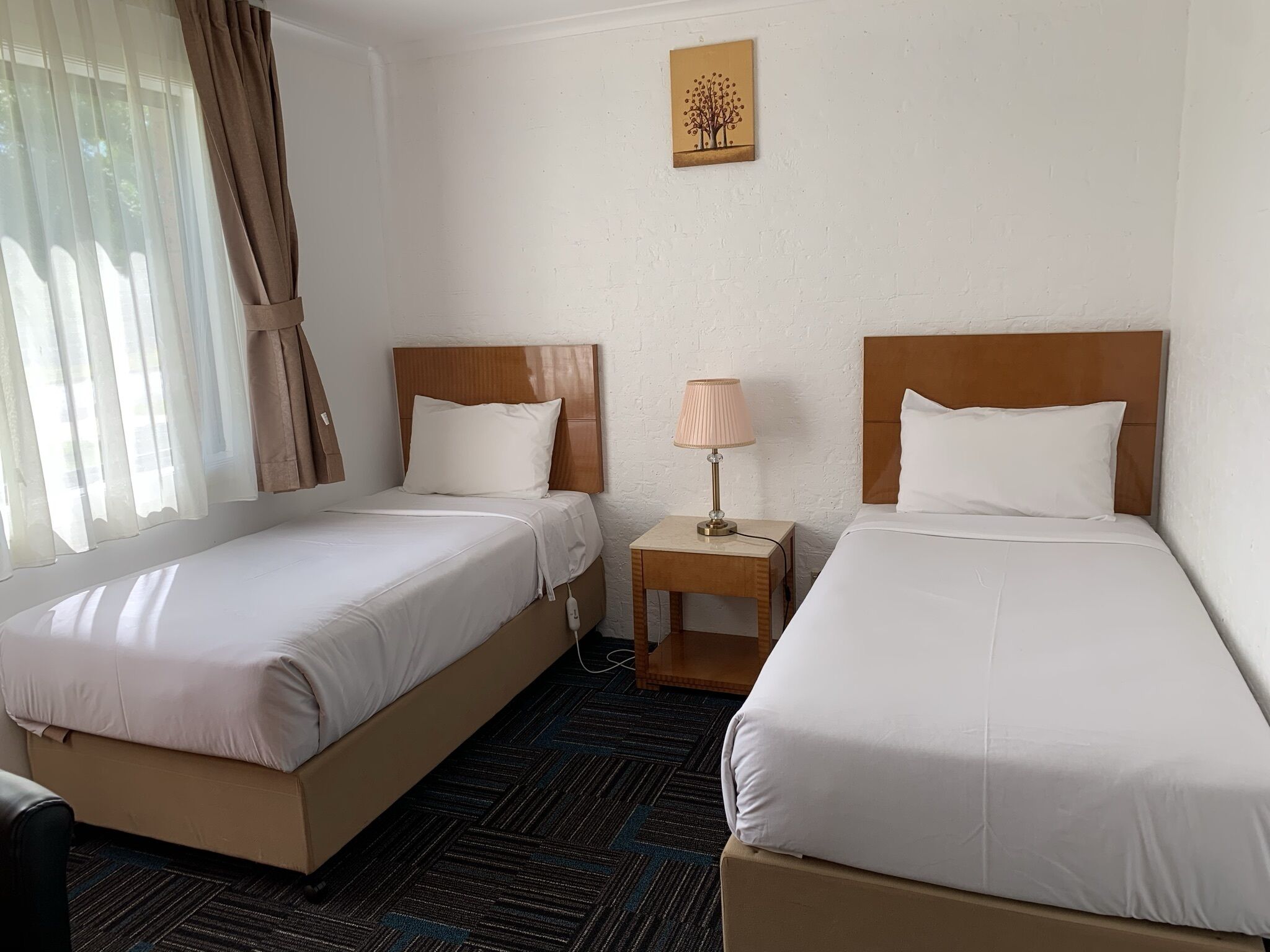 A hotel room with two single beds, a small bedside table with a lamp, a painting on the wall, and a window with curtains.