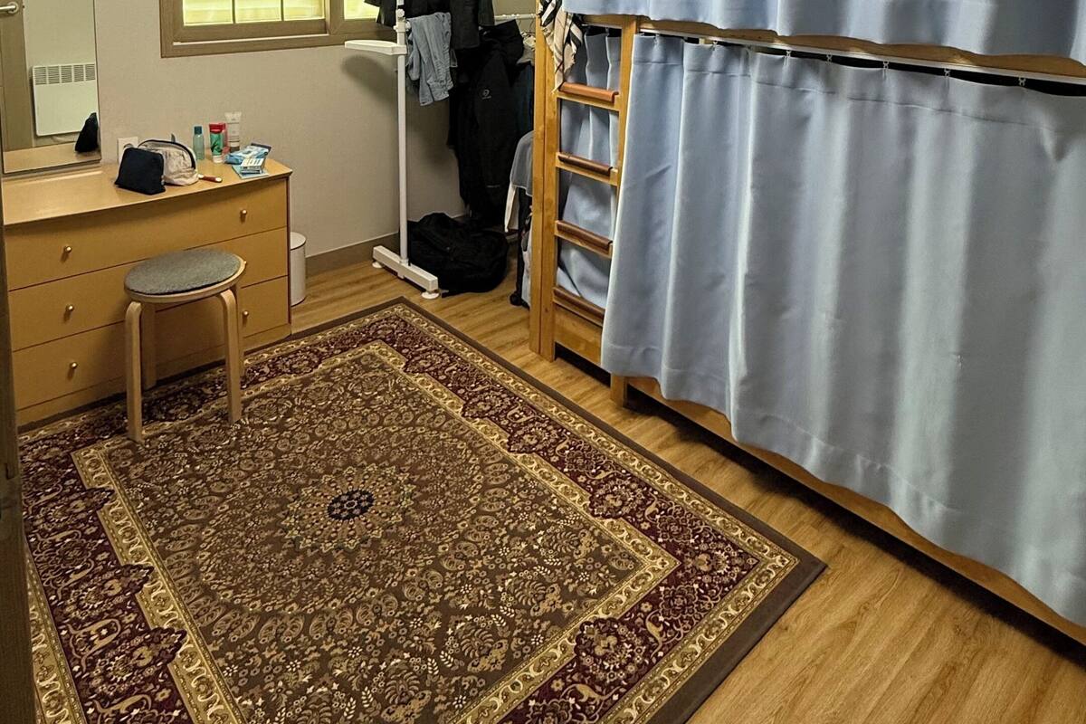 Une pièce avec un plancher en bois, un bureau avec une chaise, une grande fenêtre avec des stores, une armoire et un tapis à motifs.