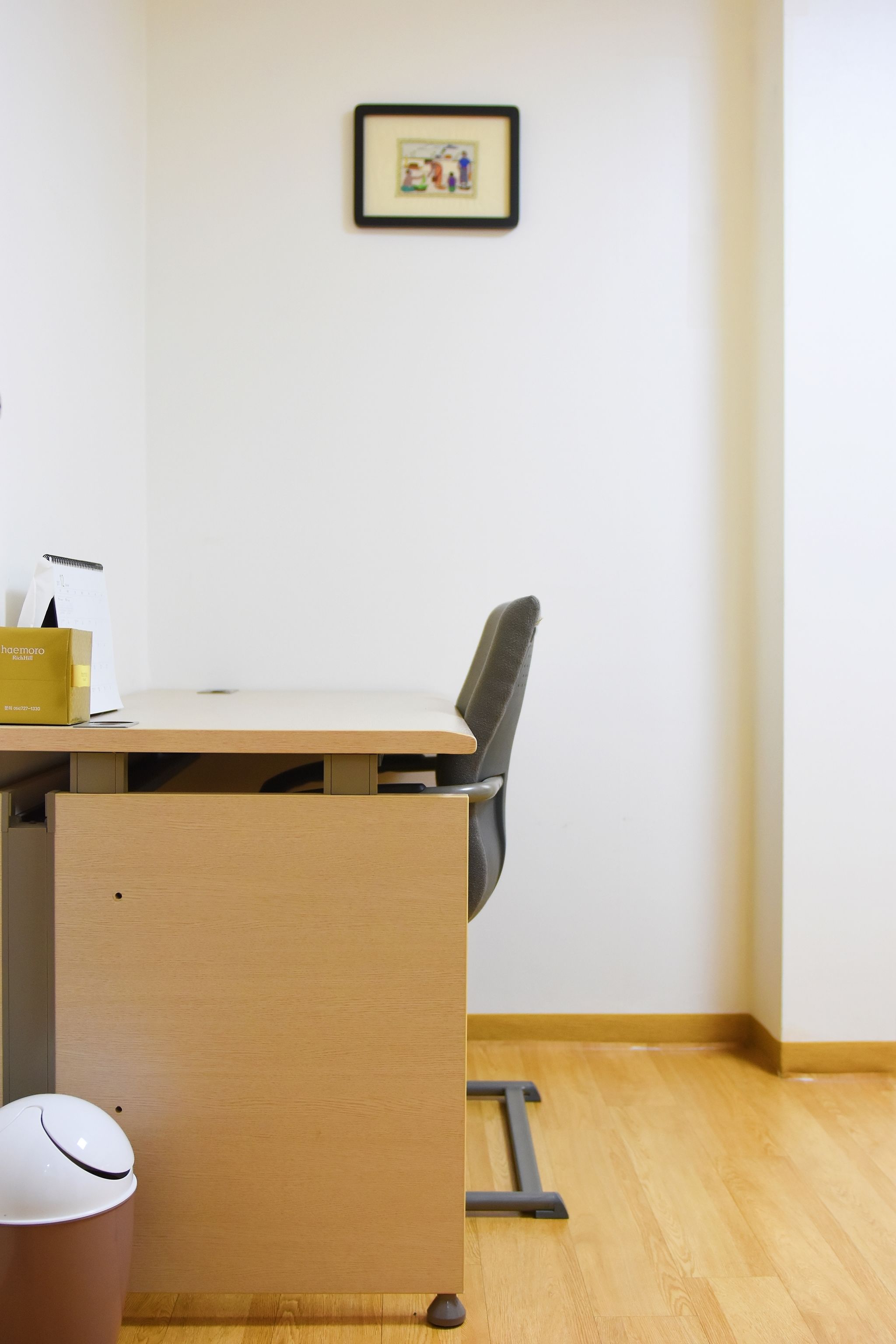 A minimalist office space with a wooden desk, a chair, a trash bin, and a framed picture on the wall.