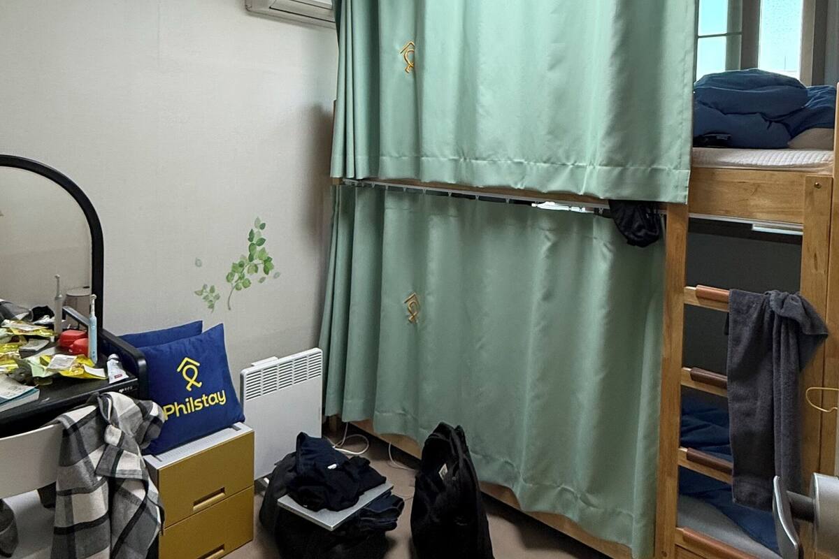 Une chambre de dortoir avec des lits superposés, un bureau avec une chaise, un miroir, un oreiller Philstay, une valise et un radiateur.