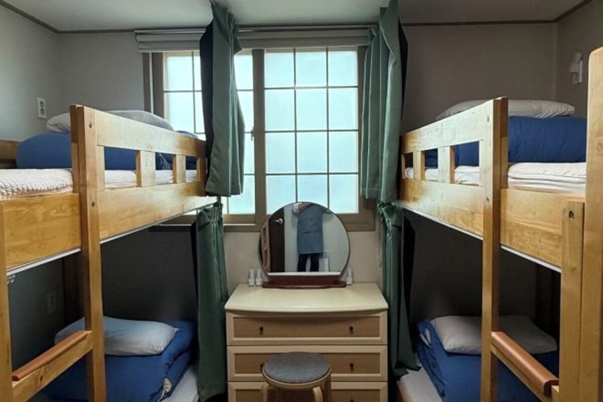 Een studentenkamer met stapelbedden, een klein kaptafeltje met spiegel en een raam met gordijnen.