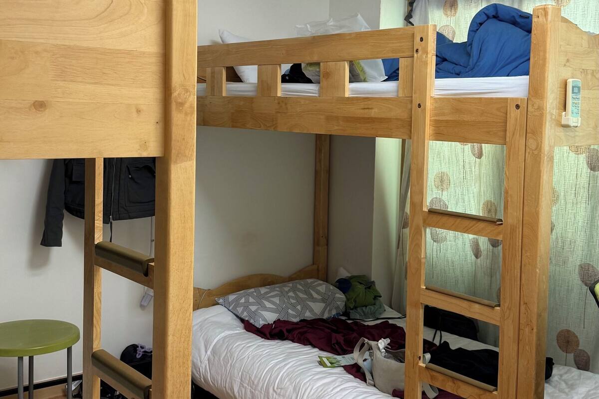 Une chambre avec un lit superposé, une tête de lit en bois, un tabouret vert et un ventilateur de plafond.