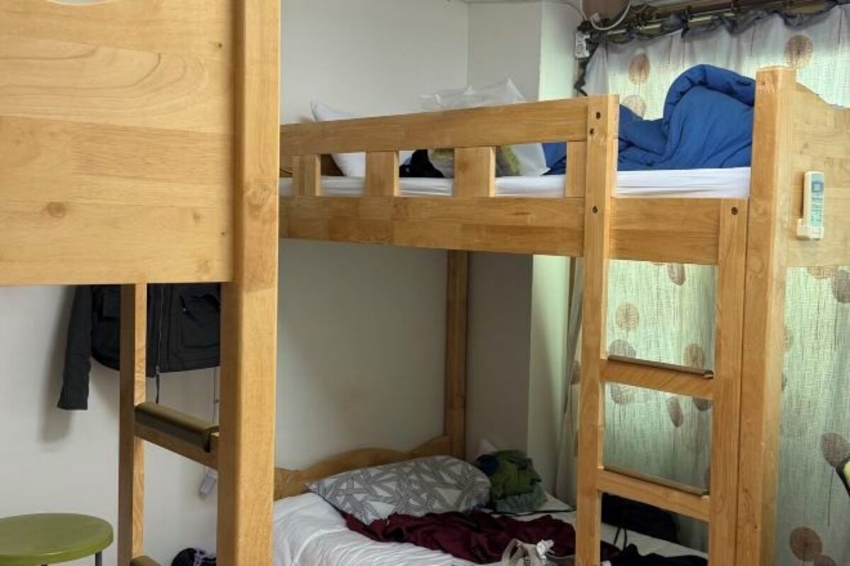 Een stapelbed met houten frame, een groene kruk en een gordijn met bladmotief.