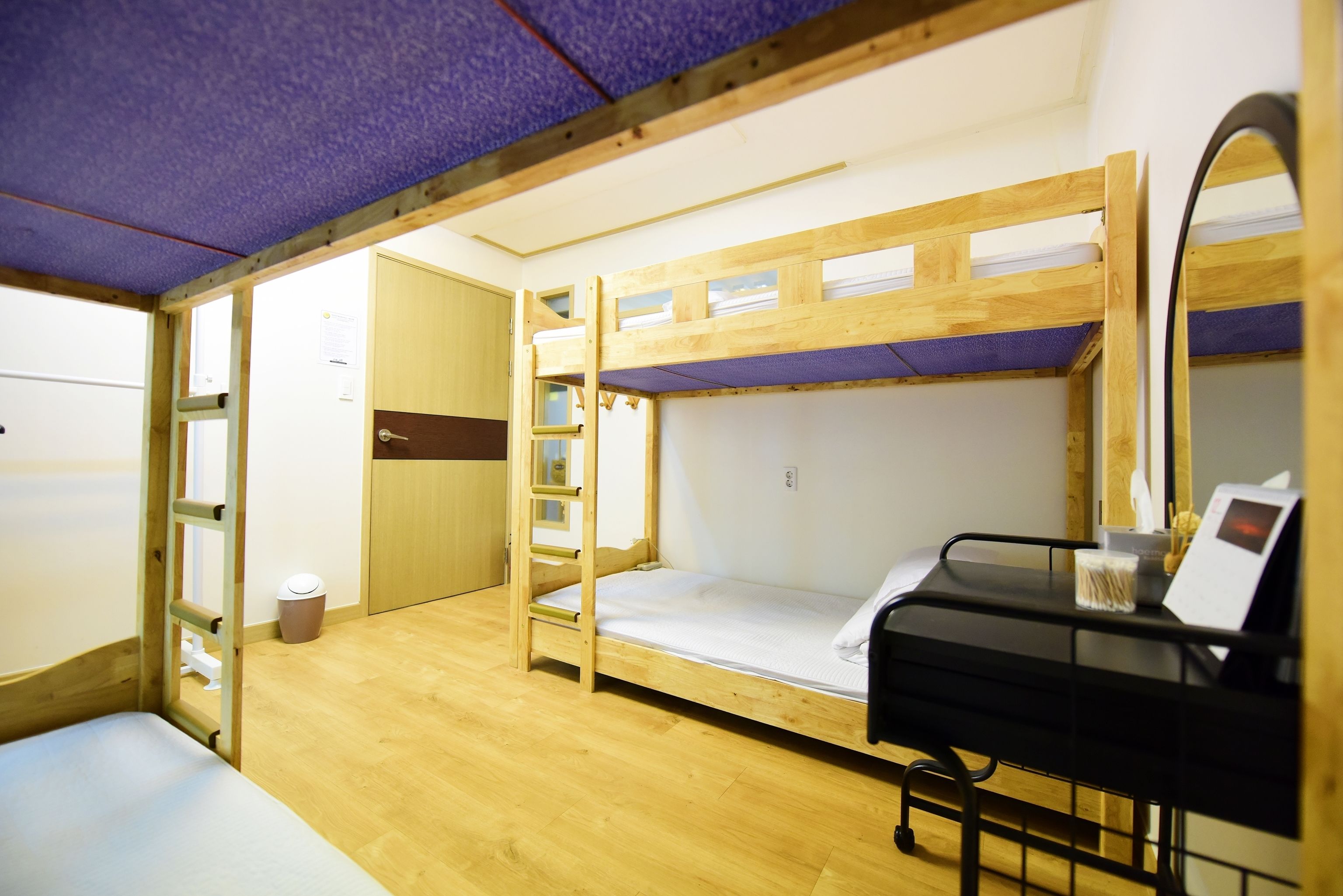 Quarto com beliche, uma escada de madeira, uma pequena escrivaninha com espelho e um cesto de lixo.