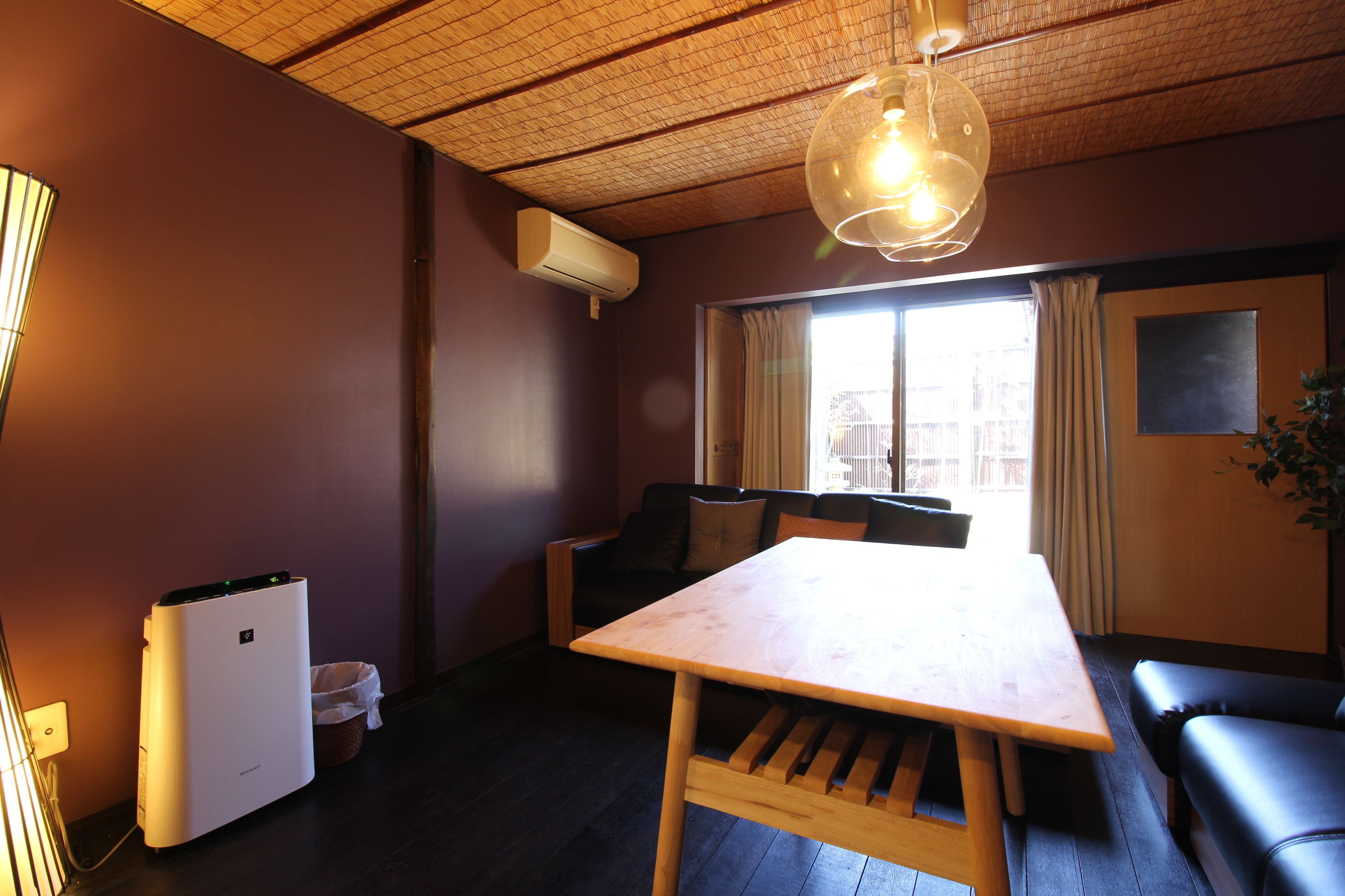 Una habitación con una mesa de madera, un sofá, un ventilador de techo y una ventana con cortinas.