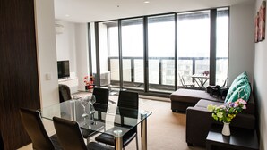 A modern living room with a glass dining table, black chairs, a sofa, and a large window offering a city view. - Madison Apartment (Melbourne)