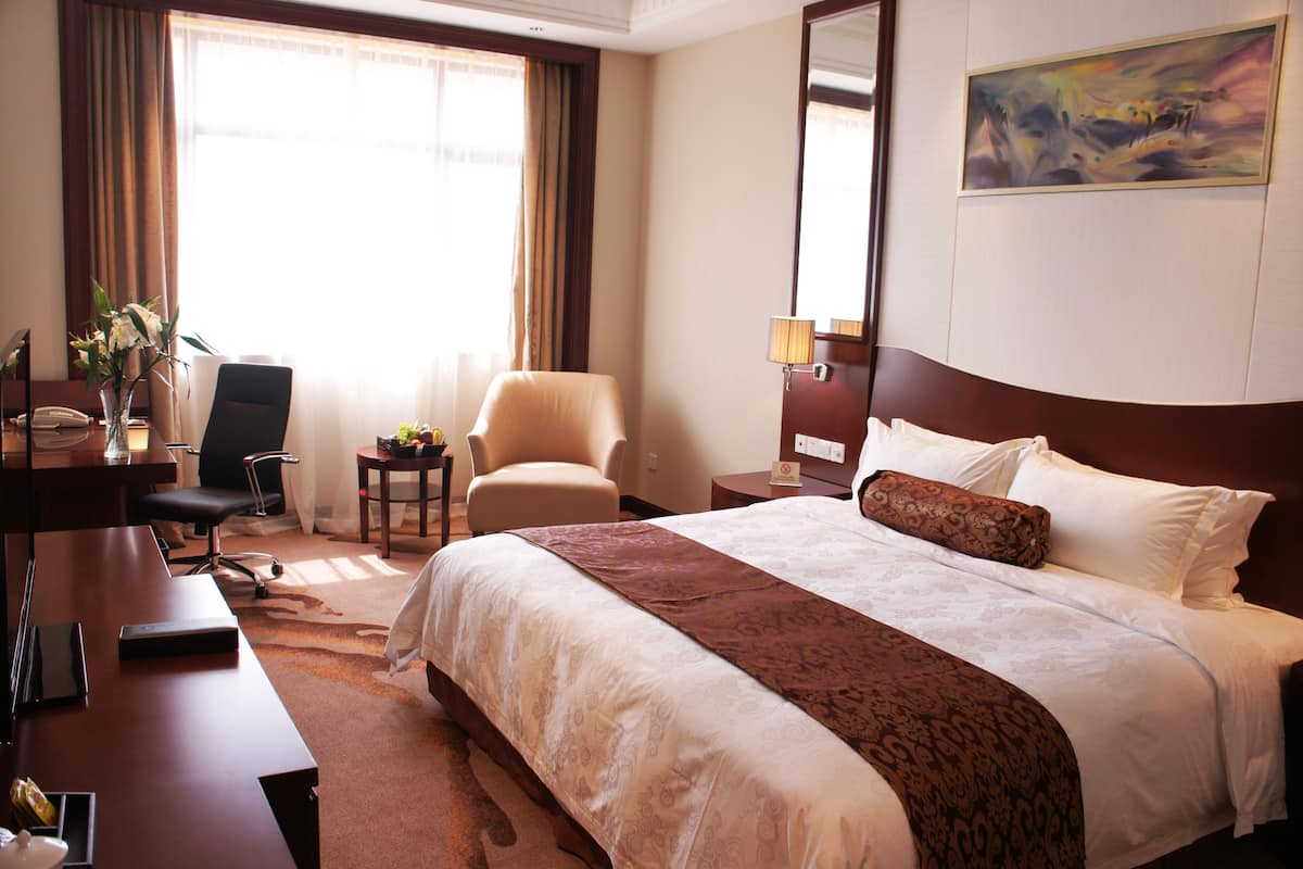 Ein Hotelzimmer mit einem großen Bett, einem Schreibtisch mit Stuhl, einem kleinen Tisch mit Pflanze und einem Gemälde an der Wand.