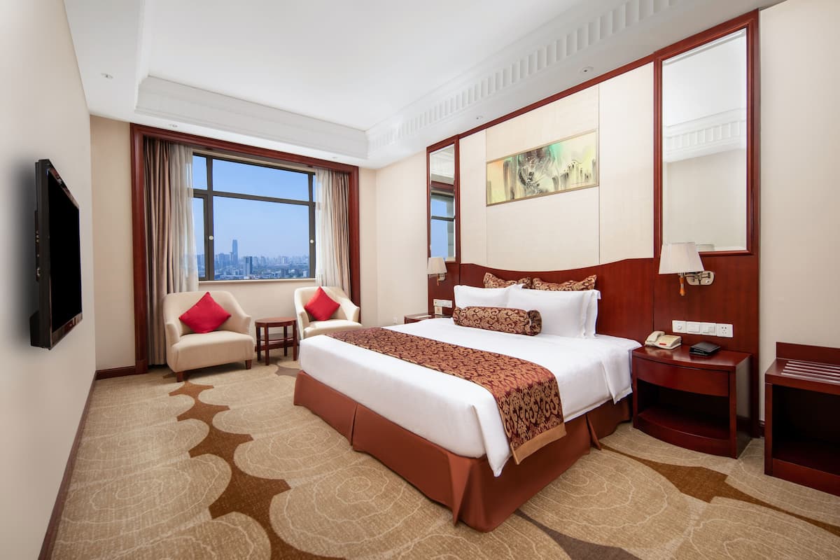 Ein Hotelzimmer mit einem großen Bett, einem Fernseher, einem Schreibtisch und Blick auf die Stadt.