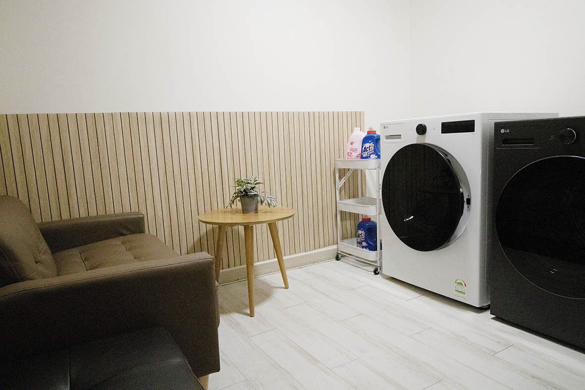 A room with a sofa, a small round table with a plant, a washing machine, and a shelf with cleaning supplies.
