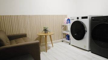 A room with a sofa, a small round table with a plant, a washing machine, and a shelf with cleaning supplies.