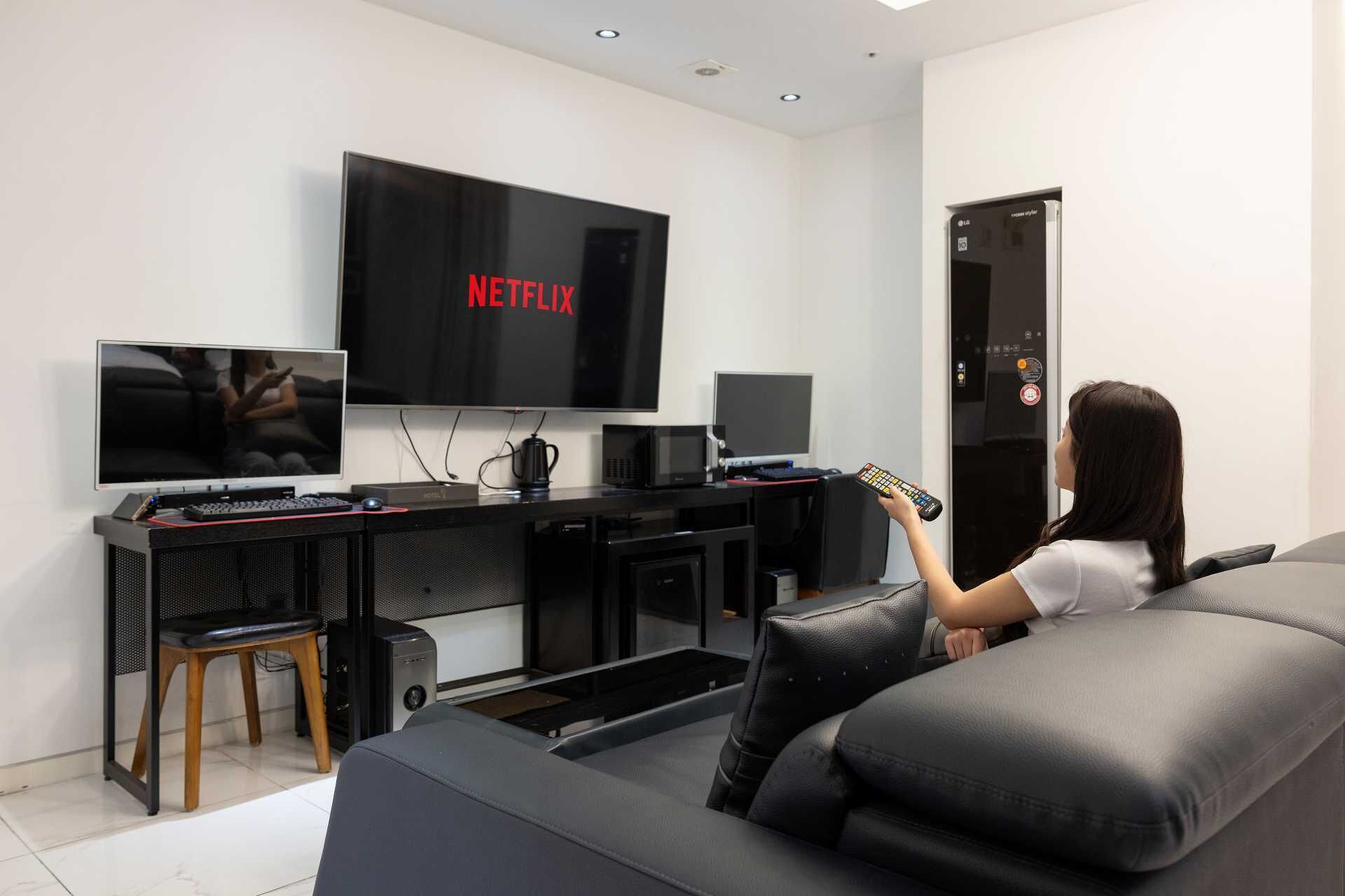 A modern living room with a large flat-screen TV mounted on the wall, a gaming console, a coffee machine, and a comfortable seating area with a sofa and a chair.