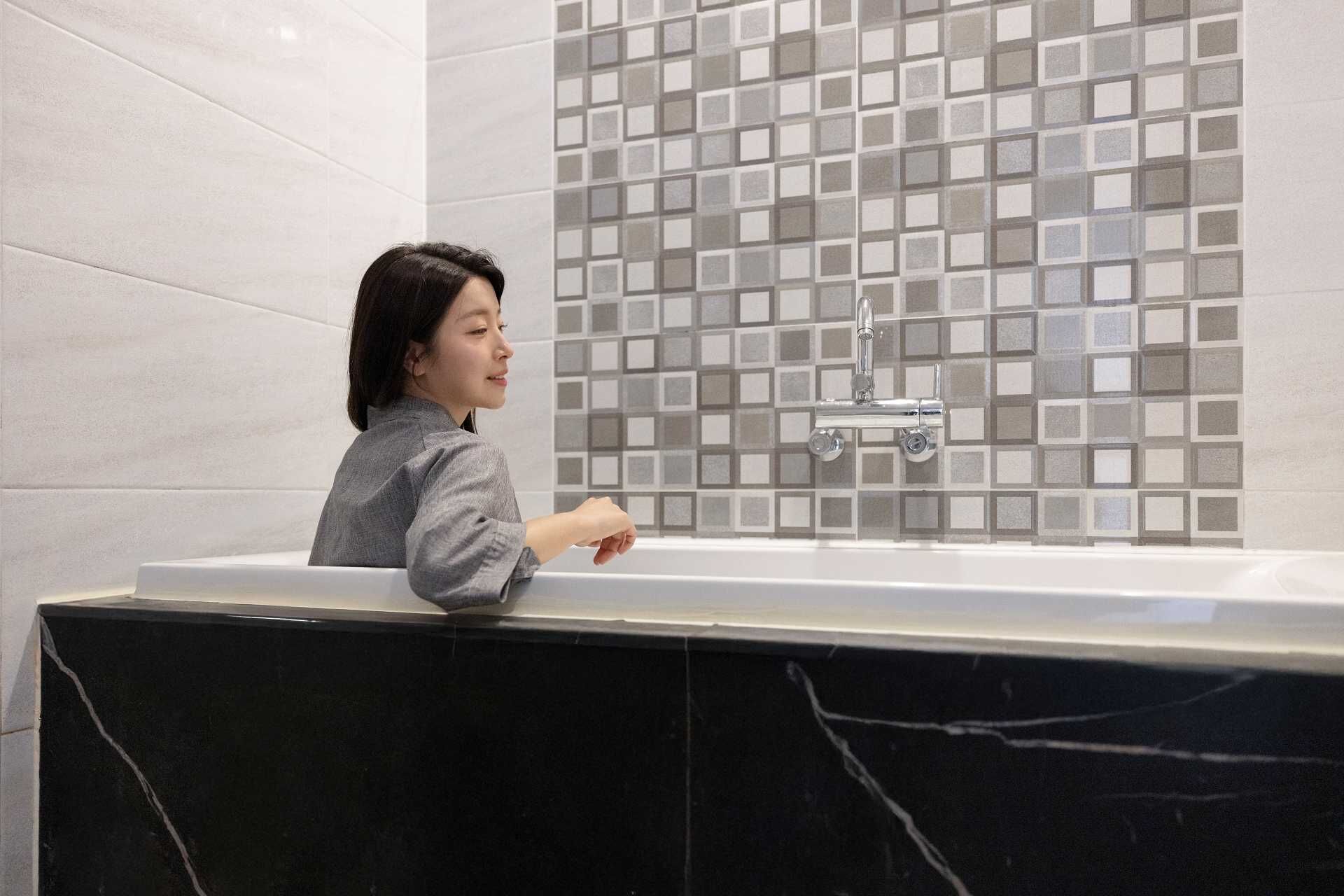 Una persona en un baño moderno con una bañera grande, paredes con azulejos y una encimera de mármol.