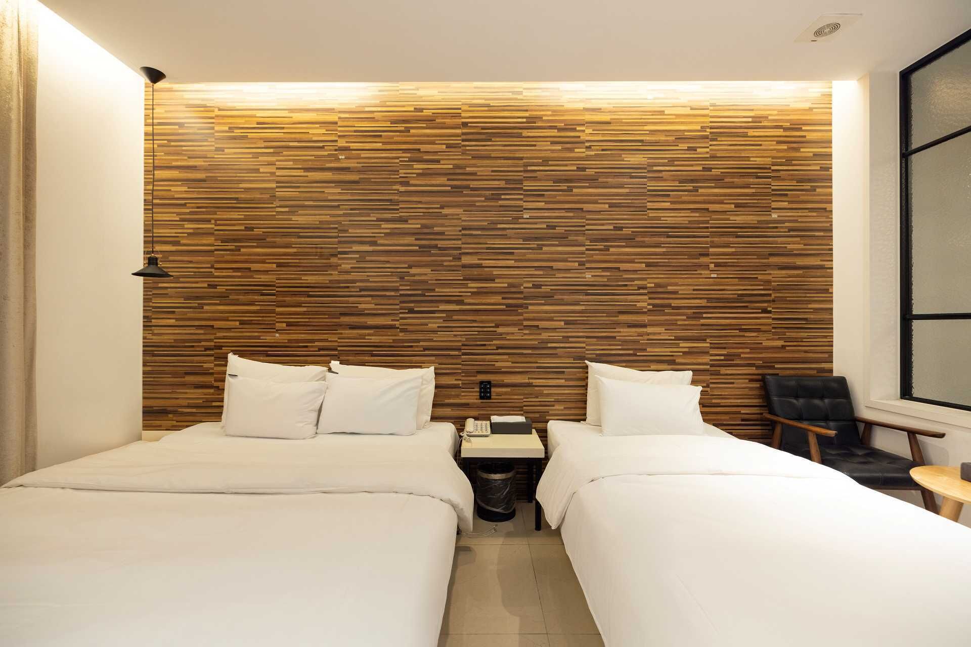 A hotel room with two beds, a wooden wall panel, a small bedside table with a phone, and a chair by the window.
