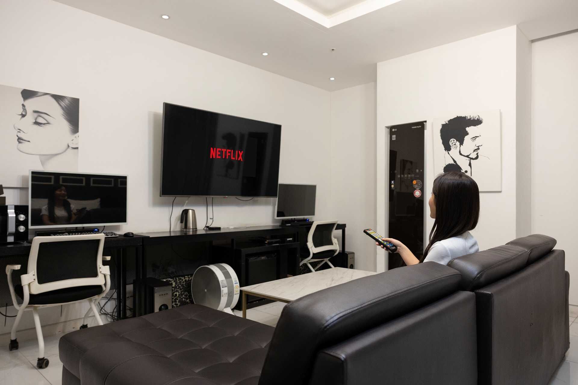 Een moderne woonkamer met een grote flatscreen-tv waarop 'NETFLIX' staat, een bank, een salontafel en een bureau met een computer en speakers.