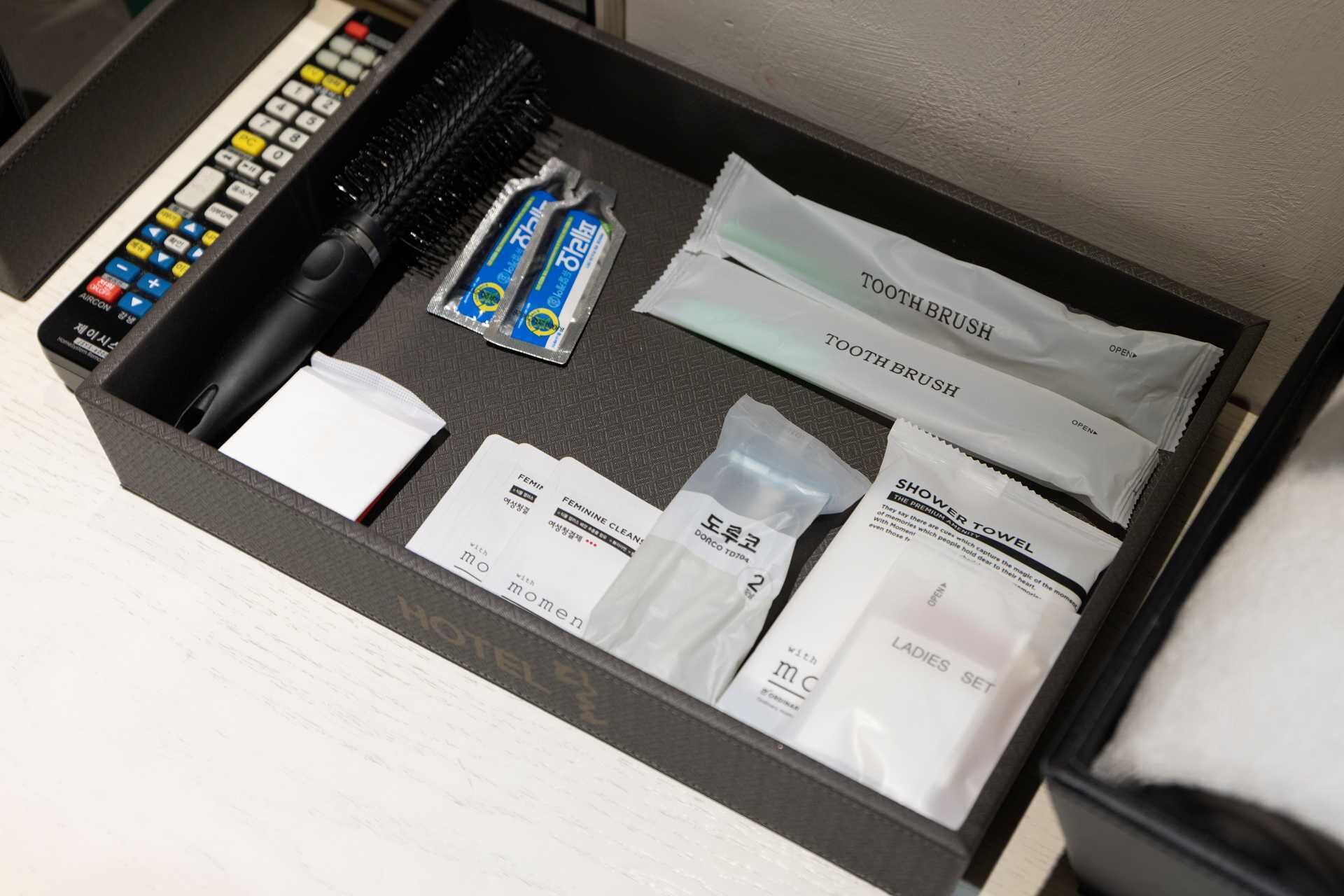 A hotel room's amenities drawer containing a toothbrush, toothpaste, a hairbrush, a towel, and a set of feminine hygiene products.