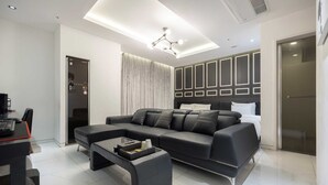 A modern hotel room with a sectional sofa, a glass coffee table, a bed with a headboard, and a wardrobe with geometric paneling.