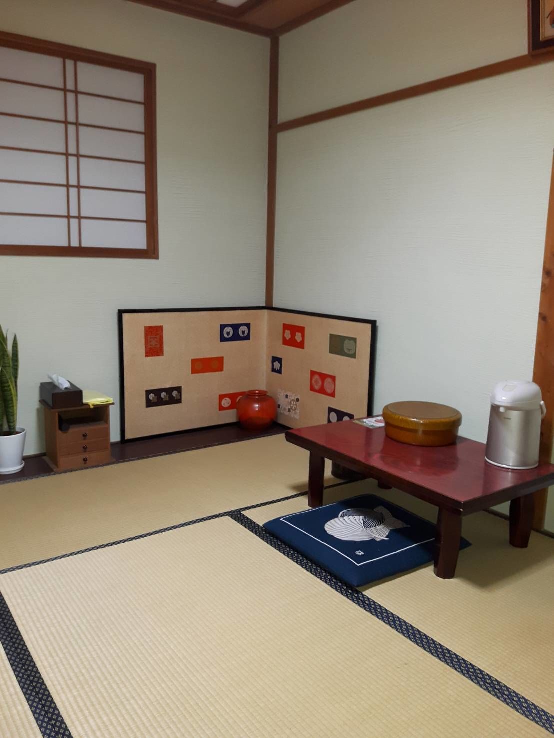 A traditional Japanese room with tatami flooring, a low wooden table, a red cushion, a small wooden cabinet, and a shoji screen with various symbols.