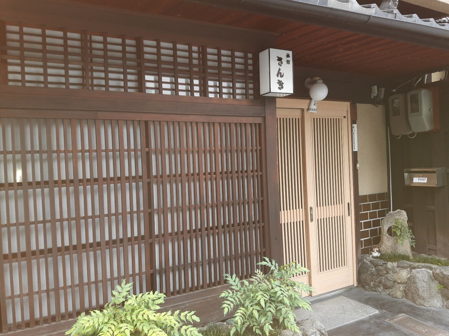A traditional Japanese gate with a wooden lattice design, a stone pathway leading to a wooden door, and a small stone lantern.