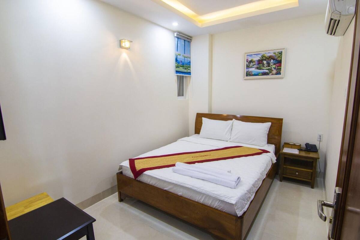 Quarto de hotel bem organizado com uma cama de madeira, uma mesa de cabeceira com abajur, uma pequena escrivaninha com cadeira e um quadro na parede.