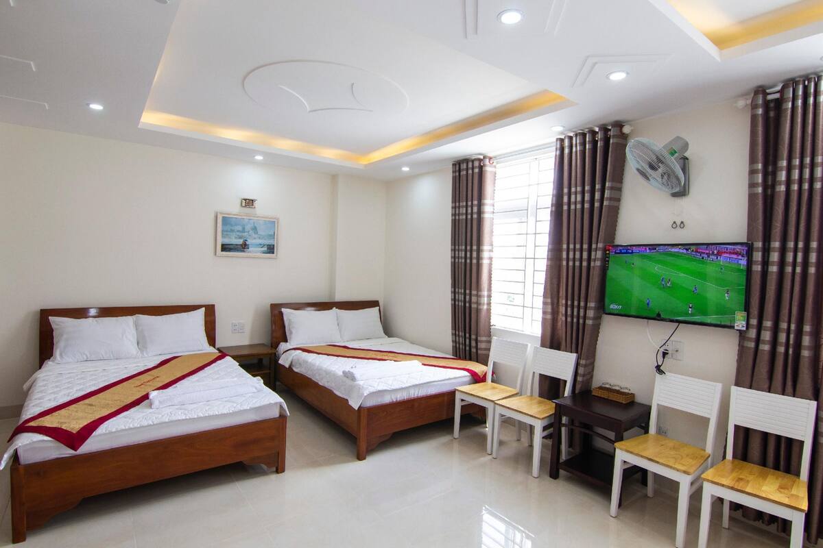 Quarto de hotel com duas camas, televisão presa à parede, ventilador de teto e janela com cortinas.
