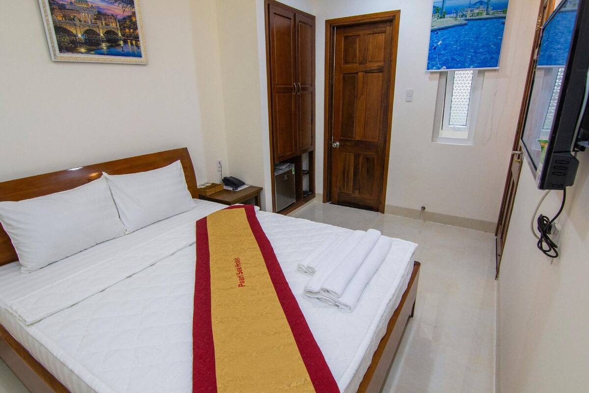 Cama bem arrumada com roupa de cama branca e toalha listrada em vermelho e amarelo, guarda-roupa de madeira, mesa de cabeceira pequena com telefone e um quadro de uma ponte na parede.