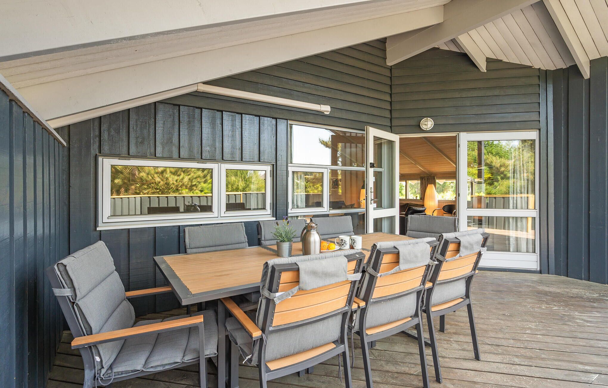 Un área de comedor al aire libre con una mesa y sillas de madera, ventanas amplias y vistas a vegetación.
