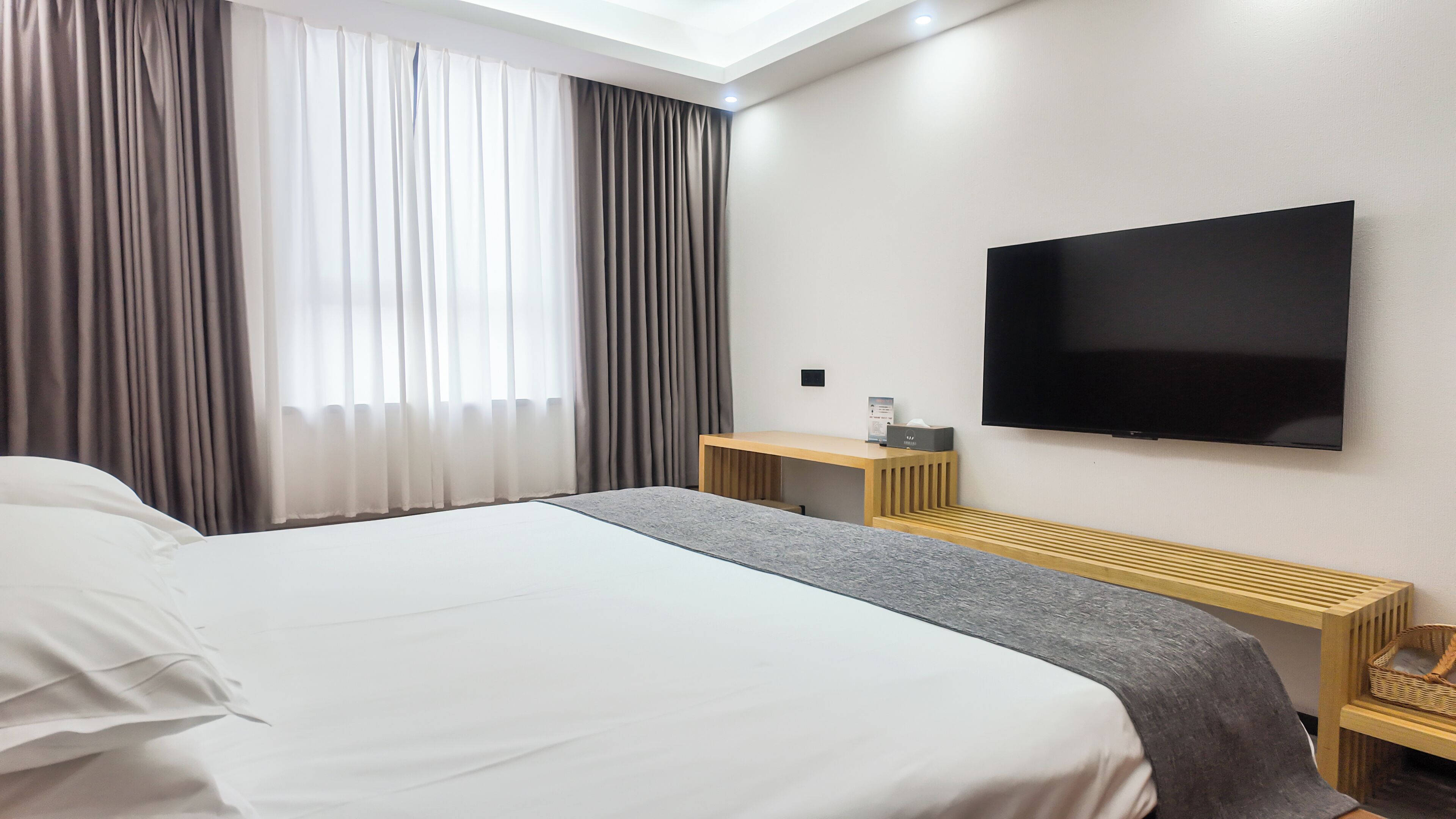 Ein Hotelzimmer mit einem großen Bett, einem Wandfernseher, einem Schreibtisch mit Stuhl und einem Vorhangfenster.
