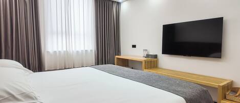 Ein Hotelzimmer mit einem großen Bett, einem Wandfernseher, einem Schreibtisch mit Stuhl und einem Vorhangfenster.