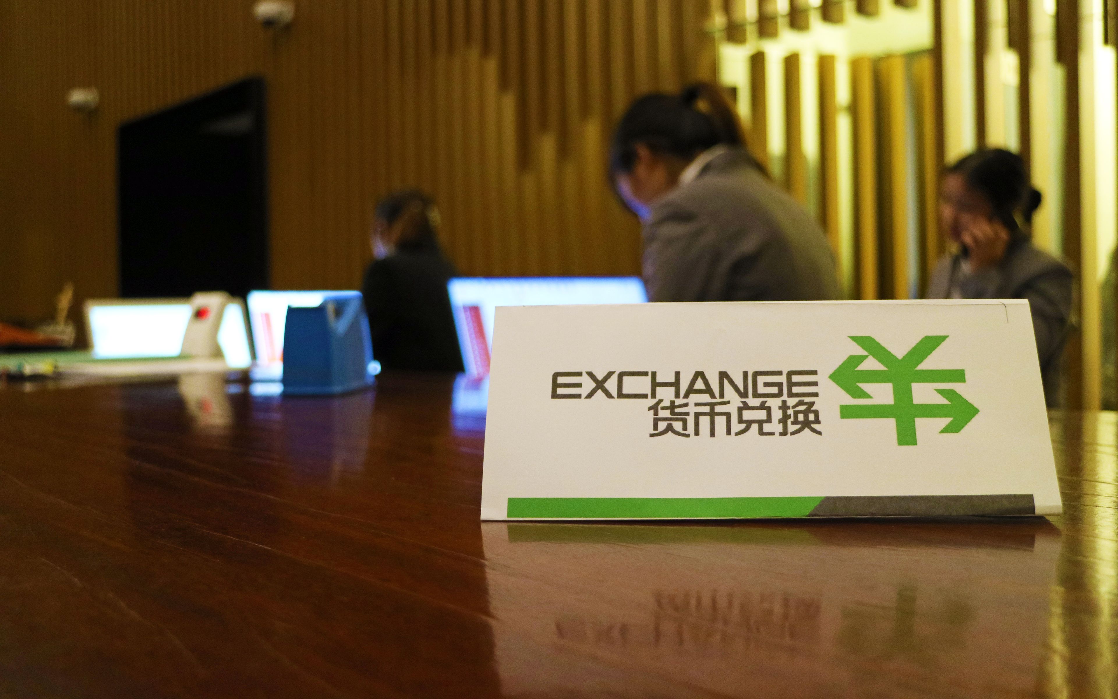 Un cartel con la palabra 'EXCHANGE' y un símbolo sobre una mesa de madera en una sala donde hay personas trabajando en computadoras.