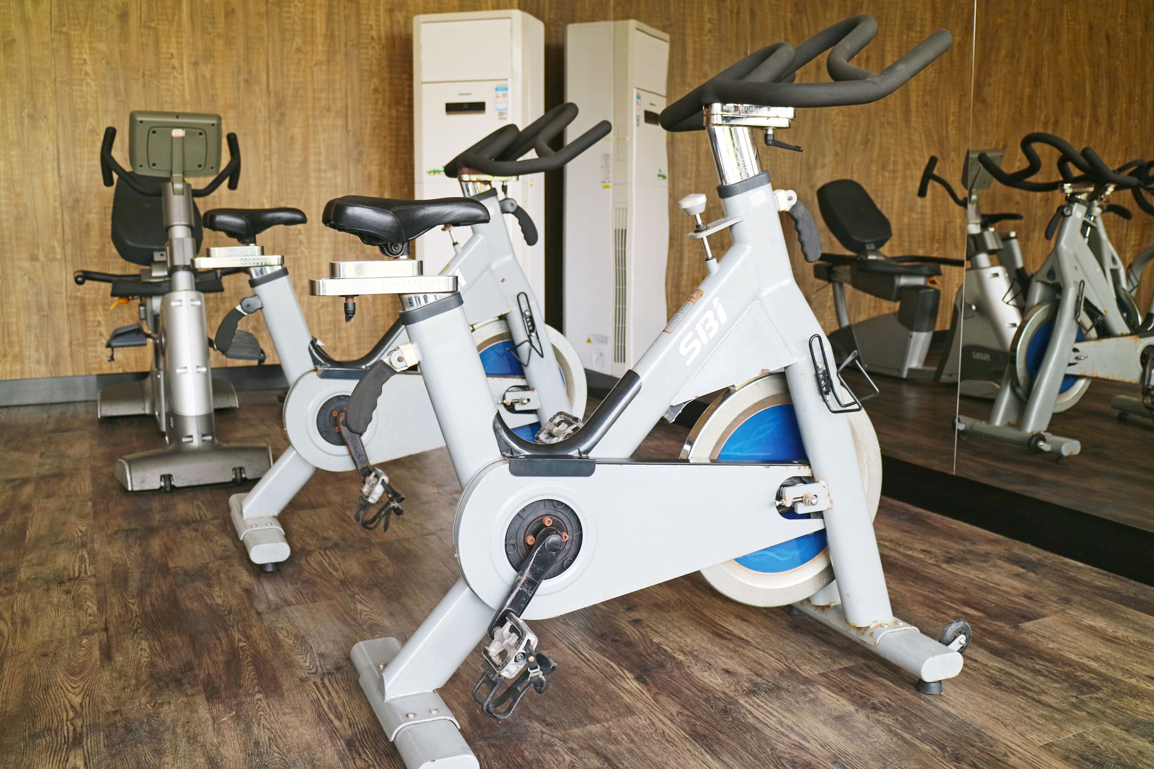 Bicicletas estáticas de gimnasio en una sala con piso de madera y un muro espejado.