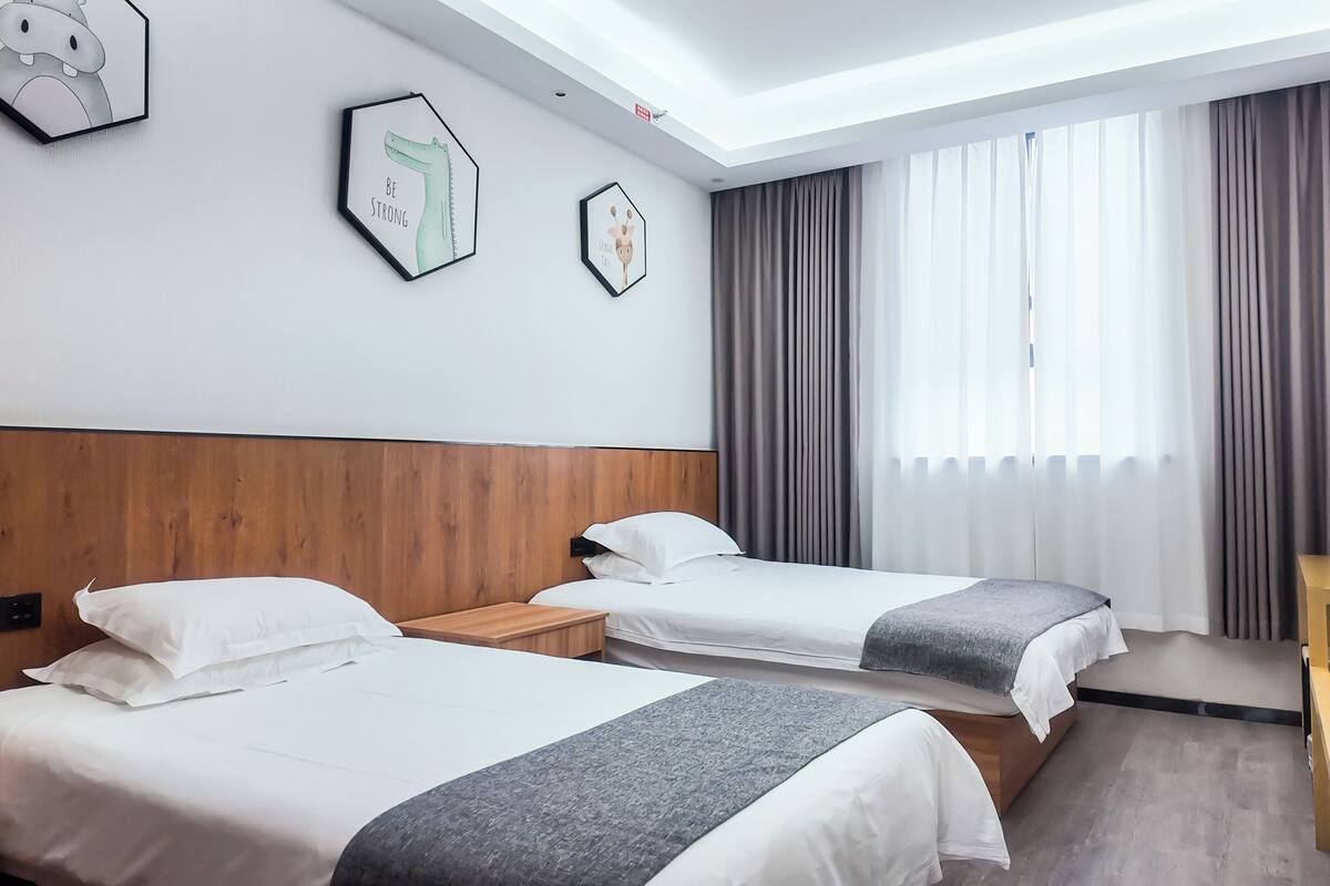 A hotel room with two beds, a wooden headboard, a desk, and a window with curtains.