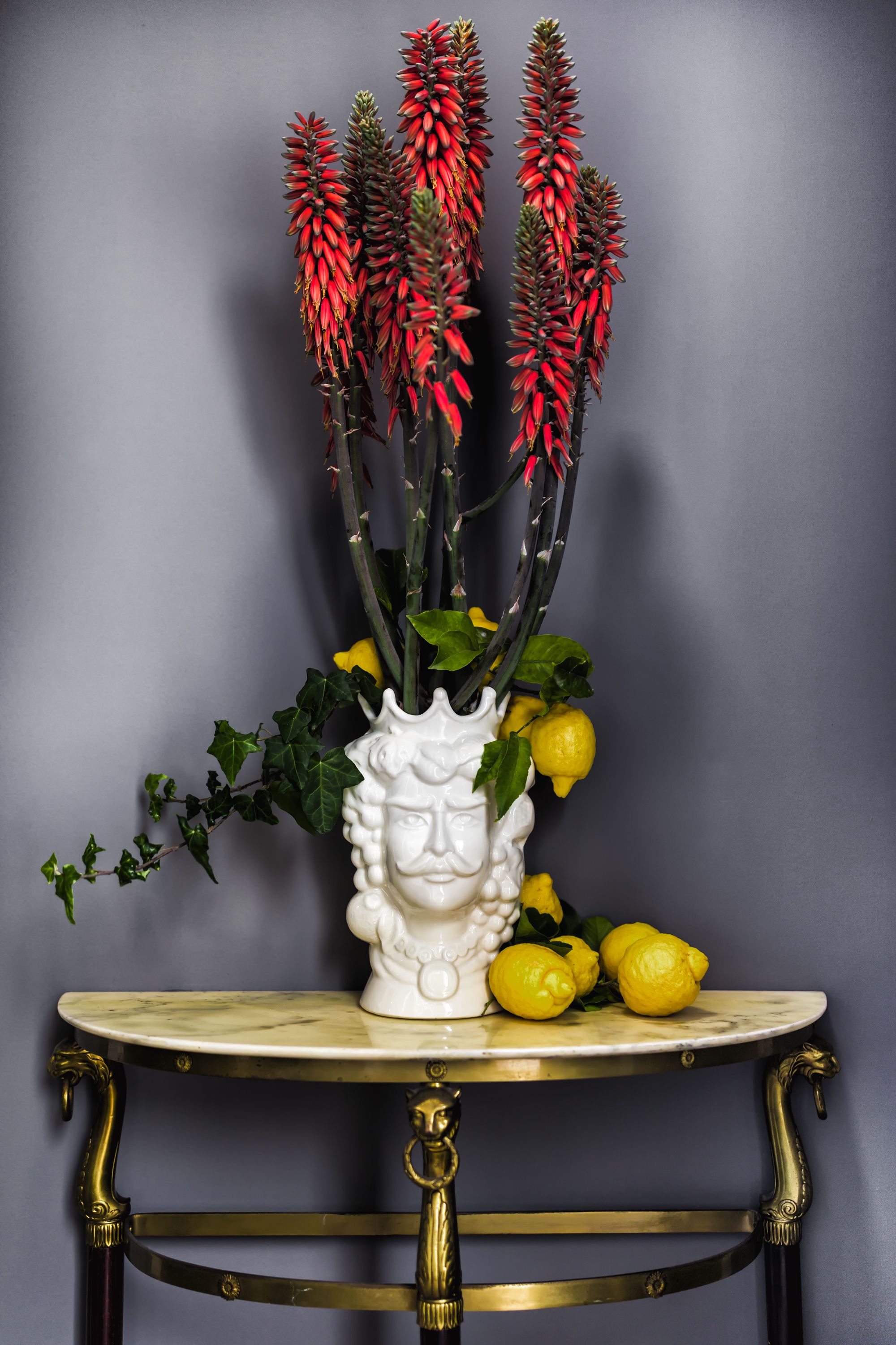 Eine weiße Vase mit Gesichtsdekor auf einem runden Tisch mit goldener Rahmen, gefüllt mit roten Blumen, gelben Zitronen und grünen Blättern.
