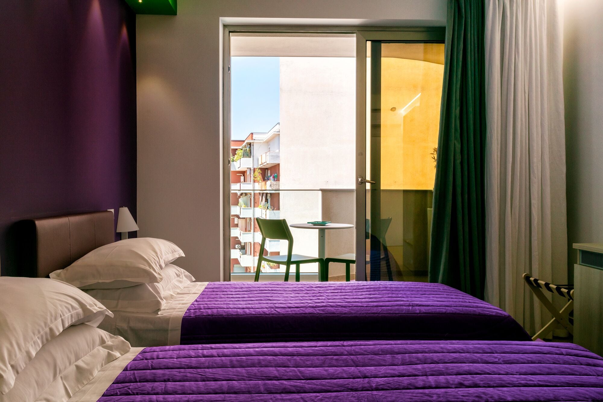 Habitación de hotel con cama, una silla y un balcón con vistas a edificios.