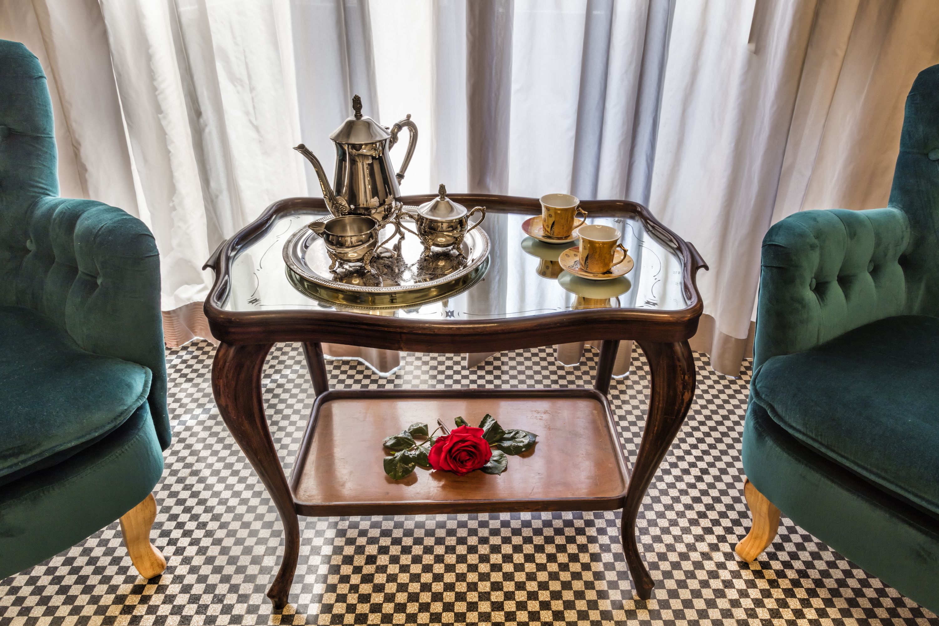 Piso xadrez, uma mesa com bandeja de talheres, uma rosa vermelha e duas cadeiras estofadas verdes.
