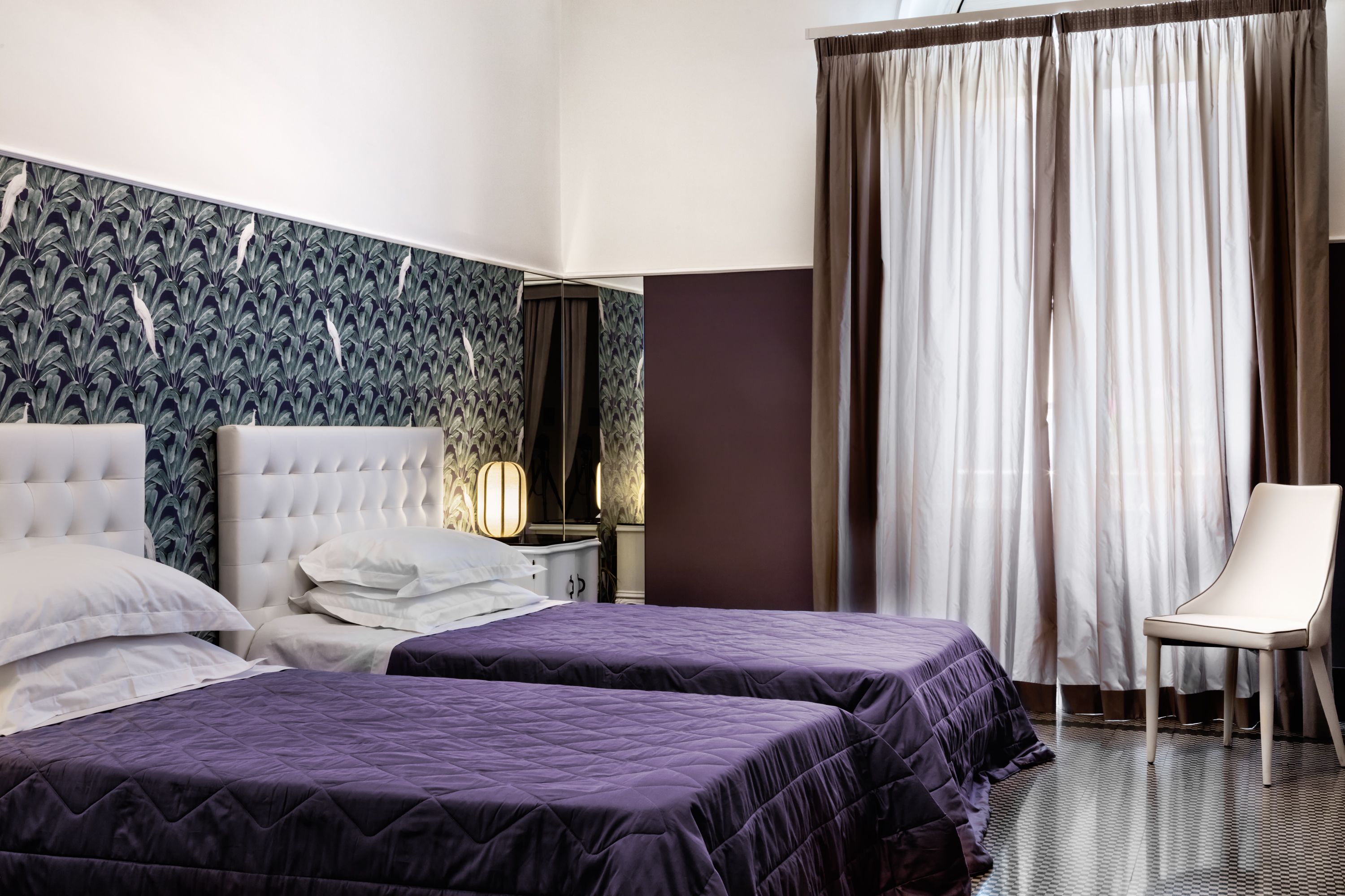 A hotel room with two beds, a chair, a nightstand, and a patterned wallpaper.