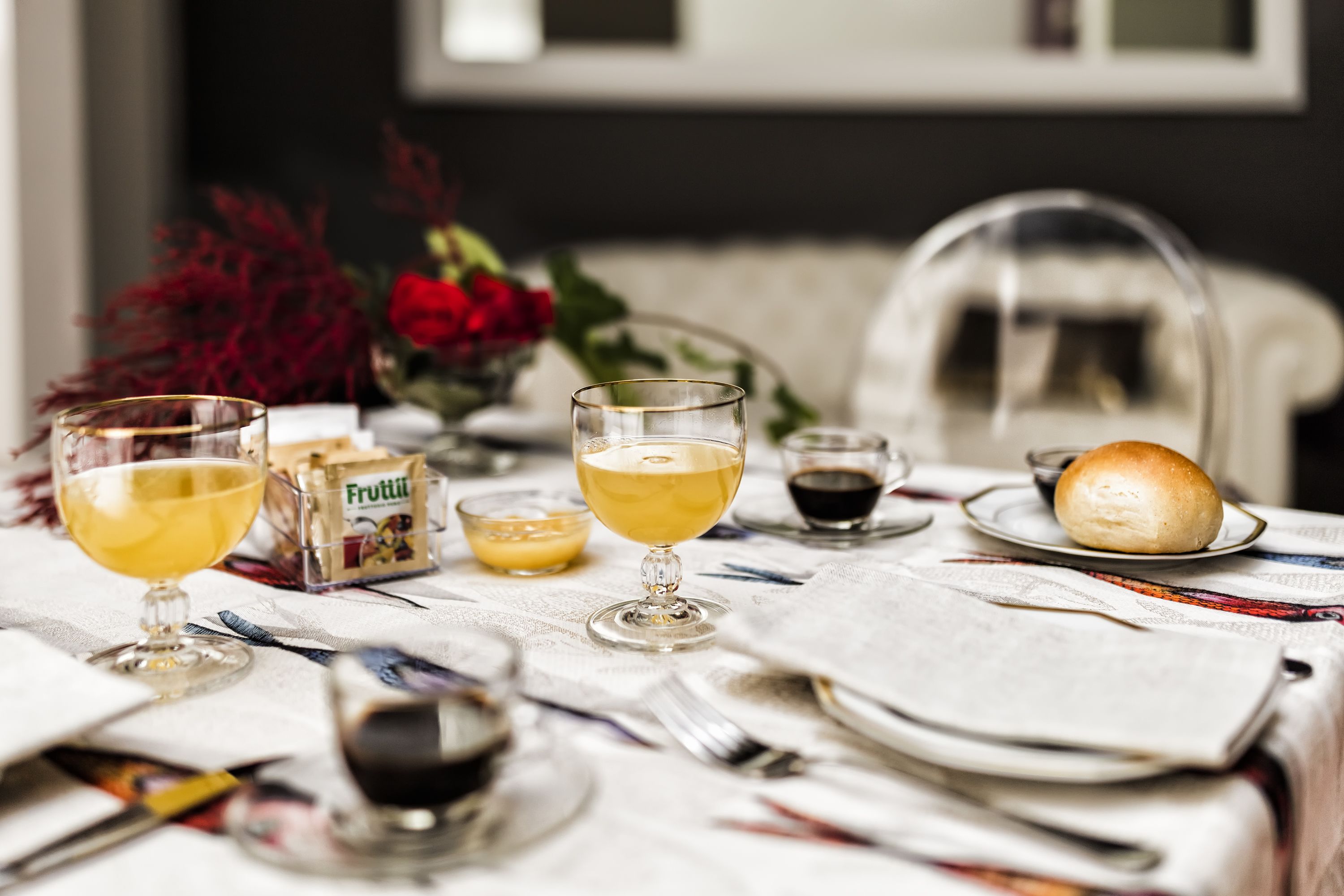 Une table dressée avec des verres remplis d’une boisson jaune, une petite boîte de fruits, un petit pain et un vase de fleurs rouges.