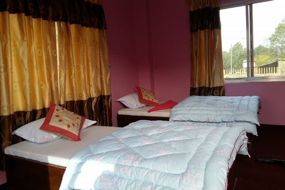 A room with two beds, each with a white mattress and a light blue quilt. There are two pillows on each bed, one white and one with a red and beige pattern. The room has a pink wall and a window with curtains.