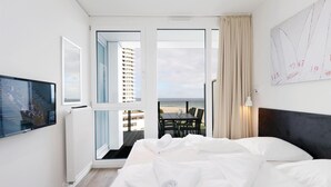 A hotel room with a bed, a TV, a balcony with a view of buildings and a pier, and a painting on the wall. - HighEnd Travemünde - Whg 603 (Lübeck Travemünde)