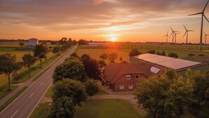 Ländliche Landschaft mit einer Straße, einem Haus mit Solaranlage und Windkraftanlagen in der Ferne.