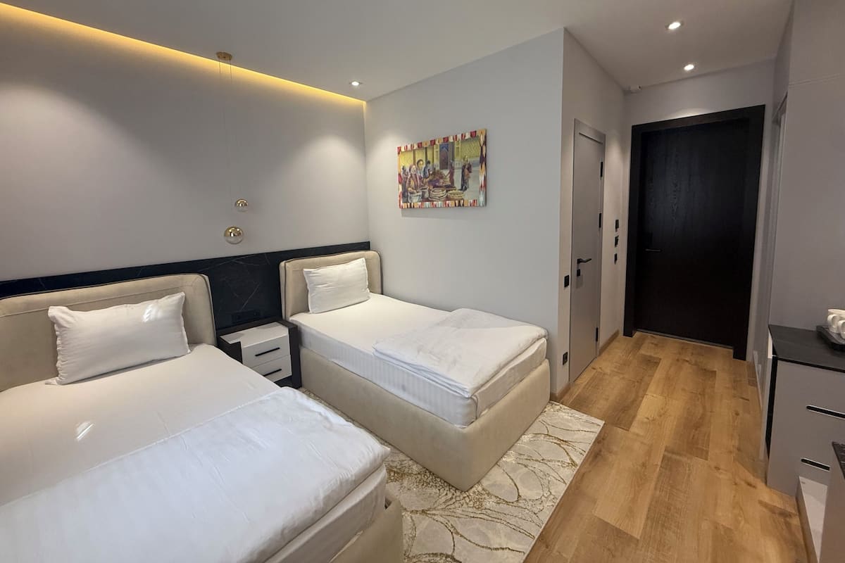 Una habitación de hotel moderna con dos camas, piso de madera, puerta oscura y un cuadro en la pared.